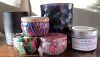 Six new candles on a surface including two large dusty black glass containers with hexagonal pattern, one small silver metal tin candle, and three colorful small round patterned candles with lids.