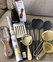 Lot of assorted kitchen utensils and tools including a pepper mill, oil dispenser, graters, strainers, ladles, slotted spatula, pasta server, and rolling pin, shown on couch.