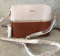 Front view of the purse showing two-tone design with light beige on top, brown on bottom, front zip pocket, and strap wrapped in plastic.