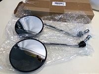 Two new round motorcycle side mirrors in chrome with threaded mounts, wrapped in plastic on a table with a cardboard box behind.