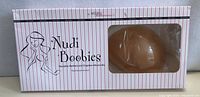 Original product packaging shows 'Nudi Boobies' brand and silicone bra visible through clear window in box.
