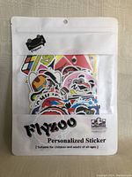 Front view of sealed white plastic pouch with clear window showing colorful stickers inside. Flyzoo logo and product name visible.