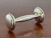 Sterling silver antique baby rattle resting on a wooden surface, showing the cylindrical handle and rounded, flared ends.