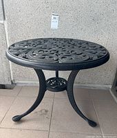 Full view of the round patio side table with three curved legs and ornate scroll work on top.