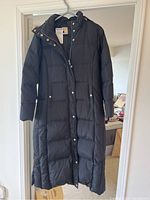 Full length view of black padded women's winter coat hanging on a hanger, showing front button and zipper closure, two front pockets, and long length.