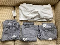 Folded white Roots zip-up hoodie with front pockets and three pairs of grey Roots sweatpants with logos, displayed on couch.