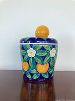Front view of the ceramic container showing painted orange fruits and white flowers with a deep blue background and orange knob lid.