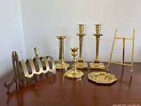 Photo showing the full lot of brass items including two pairs of candlesticks, toast holder, picture and plate stands arranged on a wooden surface.