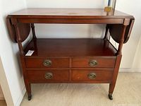 Front view of closed vintage mahogany bar cart/server on castors with two drop-leaf sides down and four drawers with brass pulls.