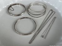 Full lot of 4 sterling silver bangle bracelets, 1 clasp bracelet, 2 necklaces, on white dish.