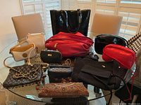 Full lot of women's handbags and purses displayed on glass table including black patent leather tote, red leather purses, black quilted bags, beige crossbody, beaded clutch, and black knitted bag