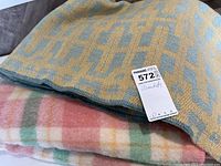 Two folded blankets stacked, top is a wool geometric patterned blanket in yellow and blue, bottom is a plaid blanket in pink, green, yellow, and white.