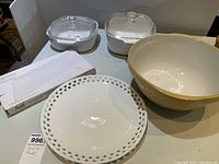 Overview of two covered Corning glass bakeware dishes, white ceramic mixing bowl, round pierced porcelain plate, and rectangular porcelain plate on table.
