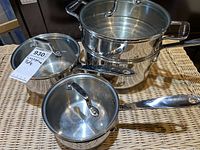 Three Lagostina stainless steel pots including one with a steamer insert and glass lids on a wicker surface. Pots have long metal handles and glass lids with metal handles.