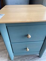 Front view showing entire small nightstand, painted matte blue with natural wood top and two drawers with round wooden knobs.