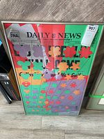 Framed colorful poster with floral patterns layered on Daily News newspaper print, leaning against wall.