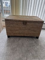 Front view of rectangular woven storage chest on carpeted floor with window blinds in background.