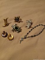 Photo showing six pieces of assorted jewelry: boot brooch, two turtle brooches, owl brooch, earrings, and bracelet with cross charm on beige surface.