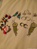 Photo showing a collection of assorted jewelry items including multiple pairs of earrings with various styles such as beaded feathers, hoops, clusters, a Santa Claus brooch, and a metallic animal-shaped item.