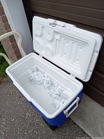 Large white and blue rectangular Coleman cooler with hinged lid open showing interior with ice packs, placed on concrete floor.