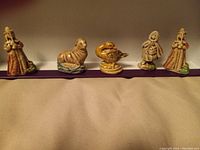Six Wade Red Rose Tea ceramic figurines arranged in a row: two robed figures with hats on each end, a seal, a bird, a woman in a cloak and bonnet, and another robed figure, displayed on a beige surface with a purple strip behind.