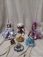 Group photo of six dolls including two porcelain dolls, a small teddy bear, and three smaller dolls in colorful clothes.