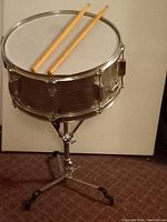 Snare drum mounted on adjustable metal tripod stand with two wooden drumsticks placed on top.