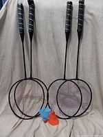 Four badminton rackets with black grips leaning against a neutral background, two plastic birdies (one blue, one orange) placed in front.