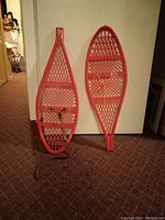 Two red plastic children's snowshoes leaning against a white surface showing shape and bindings with one snowshoe missing one strap.
