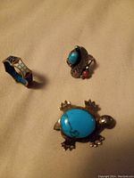 Turtle brooch with large blue stone shell, flat enamel ring, and leaf design ring with blue and red stones shown from an angle.