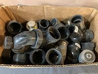 Box filled with assorted black plastic plumbing pipes, connectors, metal clamps, and valves
