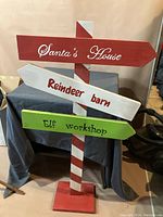 Photo of a wooden Christmas directional sign painted with red and white stripes post and three arrows labeled 'Santa's House', 'Reindeer Barn', and 'Elf Workshop'.