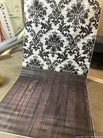 Photo showing the vinyl backdrop with a black damask pattern on white, partially rolled and wrinkled, placed on the floor in a room.