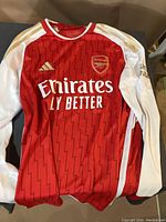 Front view of red and white Arsenal football jersey with Emirates Fly Better logo, Adidas and Arsenal logos in gold.