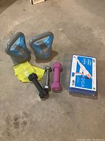 Photo showing kettlebells, dumbbells, yellow resistance band, and sealed yoga mat kit on concrete floor