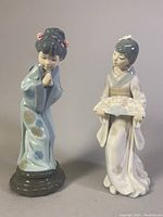 Pair of Japanese Geisha porcelain figurines shown side by side. One holds a fan, the other has hands clasped in front, showcasing their detailed traditional kimonos and hairstyles.