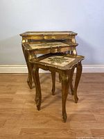 Three vintage Italian Florentine hand-painted wooden nesting tables stacked together against a plain wall on wooden floor.
