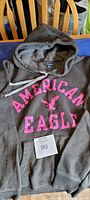 Grey American Eagle hoodie sweatshirt laid flat on a surface showing the pink branding text and hood with drawstring.
