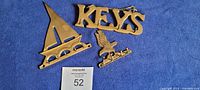 Three brass key holders laid out flat on a blue fabric surface, showing the sailboat, eagle, and 'KEYS' designs.