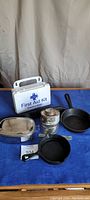 Photo showing two cast iron frying pans, First Aid Kit box, two fuel cans, stainless steel container, and a black plastic-handled tool arranged on a blue surface
