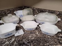 Photo showing multiple Corningware dishes with blue cornflower pattern, some with glass lids and two white plastic lids on a marble countertop.