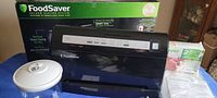 Foodsaver V3460 vacuum sealer next to its original box and packaging, showing the front of the device and included accessories like the removable drip tray.
