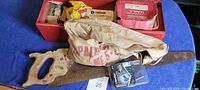 View inside the toolbox showing a hand saw with wooden handle, a canvas nail pouch, and various boxes of nails and staples.