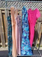 Five gowns displayed on hangers outdoors against a wooden fence, colors include peach, pink, blue floral, and blue patterned.