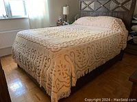 Queen size bedspread fully covering a bed with textured floral and abstract pattern in taupe tones, showing fringed edges.