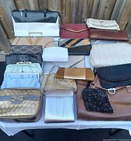 Photo showing a mix of 20 assorted purses in different materials and colors arranged on a table outside.