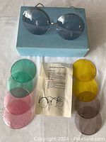 Set of round metal frame sunglasses on a blue box showing two dark grey lenses attached, six additional colored lenses arranged around, and instruction sheet visible under the lenses.
