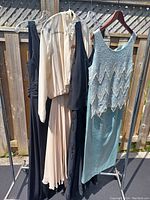Four vintage gowns and maxi dresses hanging on a clothesline outdoors, showing different styles and colors including light blue beaded dress, cream pleated gown, and two black dresses.