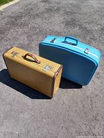Two vintage hard-sided suitcases shown outdoors on pavement: one in yellow checkered pattern with brown details, the other solid blue with silver latches.