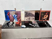 Photo showing nine vinyl LP records arranged in two rows against a pegboard background.